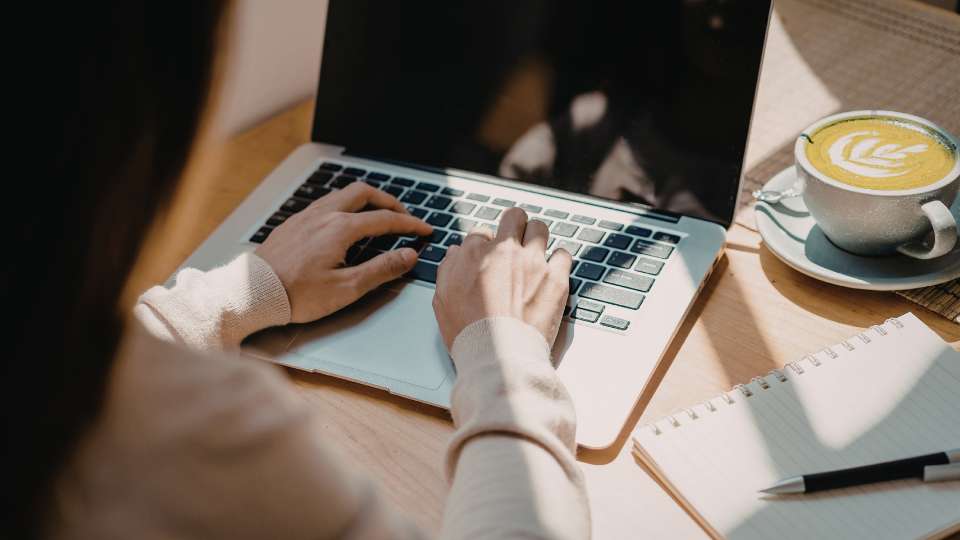 A high-angle, over-the-shoulder shot of a person typing on a laptop at a sunlit wooden desk. A cup of latte with latte art and an open spiral notebook with a pen sit beside the computer. This image depicts the active research process, illustrating the text's advice to manually verify each AI citation. By using official tools like Crossref to check DOI strings, researchers can uphold research papers citation rules and ensure every reference in their work is legitimate rather than a hallucinated link.