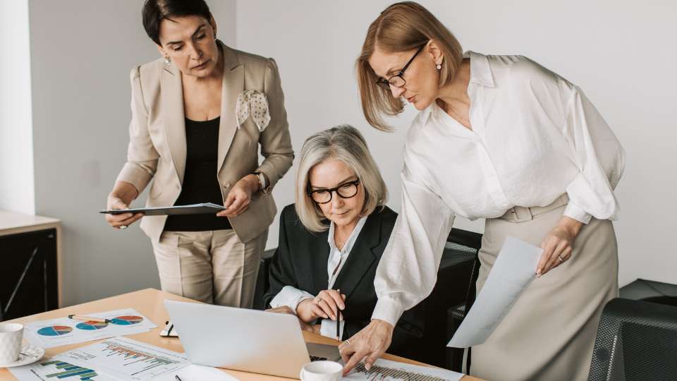 Three researchers—representing the diverse expertise of a global team—collaborate intensely over a laptop and data charts. This image illustrates the "Potluck Dilemma" discussed in the text: while one researcher provides the data analysis and others provide the documentation and oversight, the lack of a formal intellectual property agreement among them creates a tense atmosphere of scrutiny as they attempt to finalize their breakthrough results.
