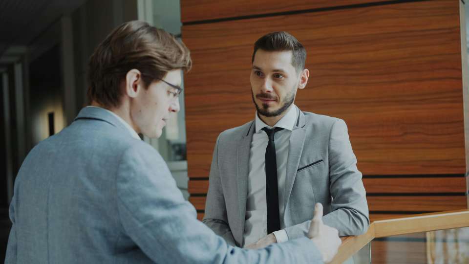 Two male researchers in professional suits are engaged in a conversation in a modern office hallway. The man in the foreground, with his back to the camera, is speaking while the man facing him listens with a guarded, skeptical expression. This image represents the potential for ethical conflict and "opportunism" mentioned in the text, where blurry intellectual property boundaries can lead to tense negotiations over data access and unearned authorship demands.
