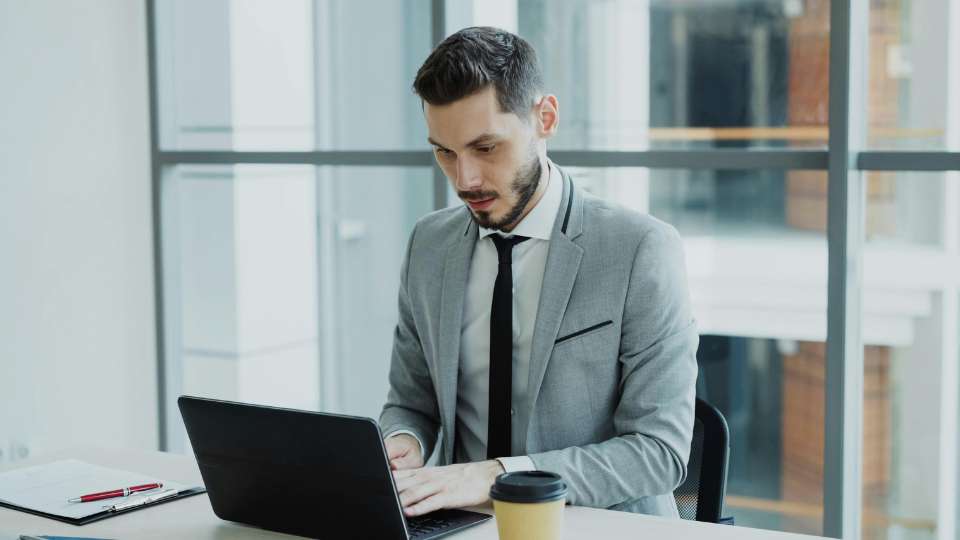 A man in a sharp grey suit and black tie sits at a desk in a modern, glass-walled office, looking down at his laptop with an expression of intense scrutiny. His focused posture suggests a meticulous review of the data on his screen. This image illustrates the "Formatting Quirk" hack, representing the level of detail a researcher must apply when reviewing an automatically generated literature review. It underscores the text's advice: while formatting errors are human, missing or inconsistent metadata from an AI is often a sign of a "blind guess" that requires immediate verification to maintain academic standards.