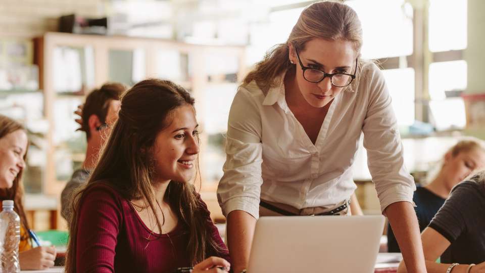 A female professor with glasses leans over a student's desk in a sunlit classroom, both looking intently at a laptop screen. The student is smiling, while the professor maintains a focused, evaluative expression, representing the "Ultimate Professor Test." This scene illustrates the necessity of human oversight in the research process. It underscores the text's final point: that while AI can assist in building a bibliography, the deep, historical knowledge of a mentor is the most reliable safeguard to fact-check an AI citation and ensure the total legitimacy of a scholarly project.