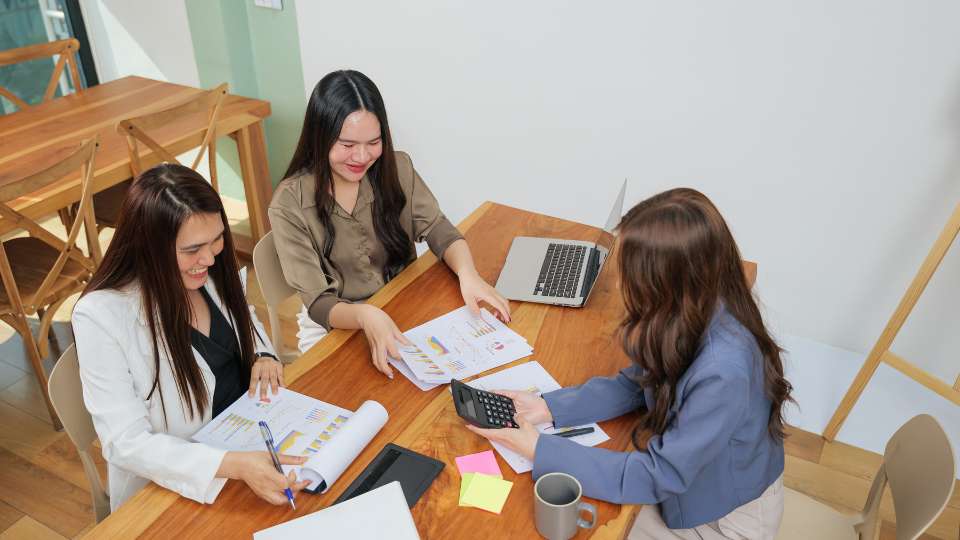 A smiling research dream team of three women collaborating around a wooden table, happily analyzing data charts together.