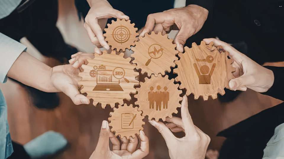 A group of people's hands are shown connecting six interlocking wooden gears of various sizes, symbolizing collaborative synergy. Each gear is engraved with a specific icon representing different business or research functions: a target for goals, a magnifying glass for analysis, a person holding a key for solutions/leadership, a group of people for teamwork, a document and pen for formal agreements, and a laptop displaying "SEO" and growth charts for digital marketing. This visual metaphor illustrates how a research team can promote your research by dividing a multi-platform strategy into manageable, interconnected parts, where each member's contribution keeps the overall promotional machine moving efficiently.