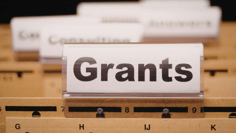 A close-up of a filing cabinet drawer with several labeled tabs. The prominent front tab reads "Grants" in bold black letters. The image symbolizes the organized approach required to secure a research grant, representing the foundational financial support needed to build a successful academic career.