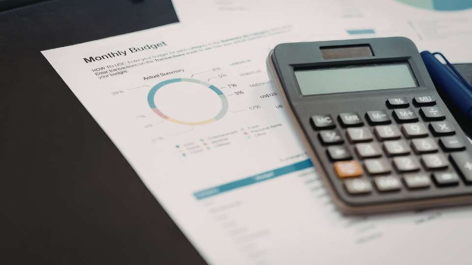 A close-up shot of a "Monthly Budget" document featuring a colorful pie chart and an "Actual Summary" table, with a calculator and blue pen resting on top. This image highlights the financial precision needed in an academic career to justify the costs of lab equipment and salaries, ensuring that every penny of a research grant is accounted for and professionally managed.