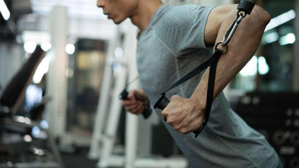 A man in a gym uses cable resistance to open his chest and shoulders. This movement illustrates the importance of stretching the torso to reverse the "hunch" caused by long hours of research, effectively mitigating researcher back pain.