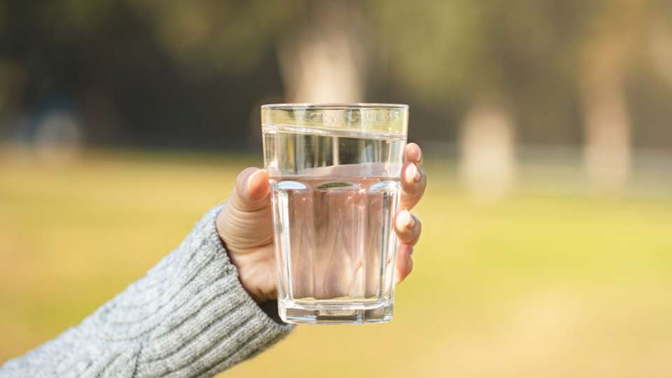 A hand holding a clear glass of water against a blurred outdoor background. This image emphasizes the importance of hydration as a core pillar of healthy meal planning to support spinal health and mitigate tech neck symptoms for digital researchers.