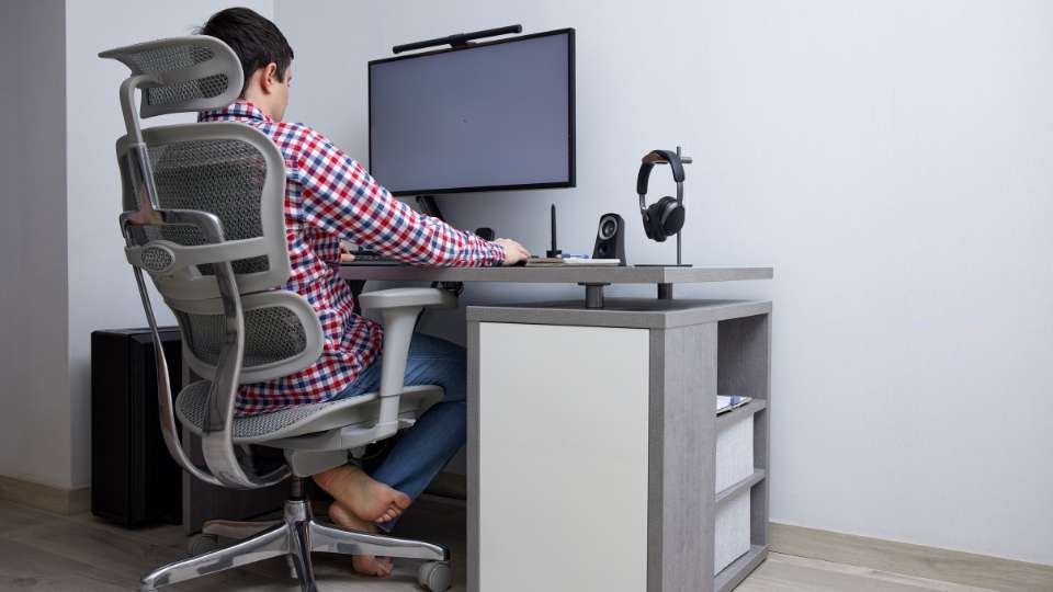 A man sits in a high-quality ergonomic chair at a desk equipped with a raised monitor, external keyboard, and mouse. This workstation setup demonstrates how to prevent tech neck symptoms and maintain productivity throughout a long academic career.