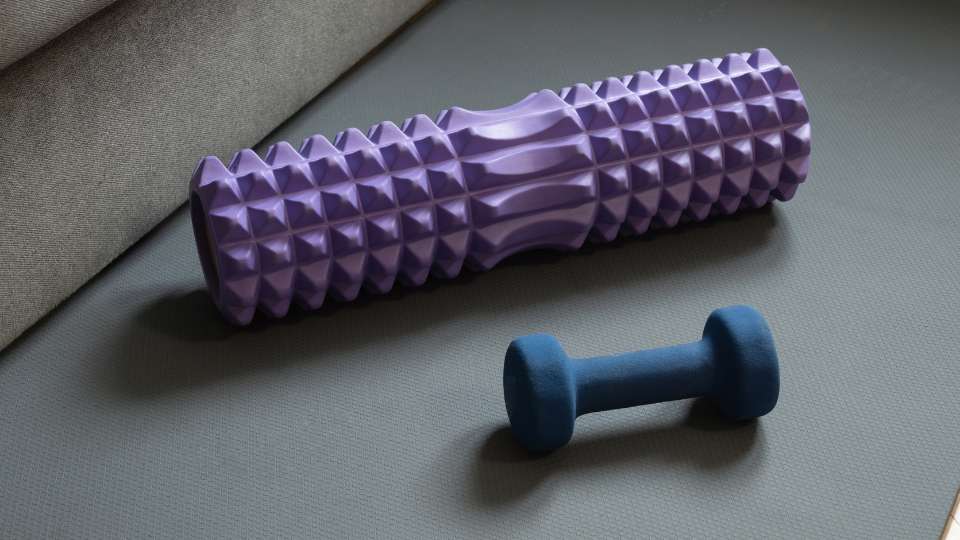 A purple textured foam roller and a blue dumbbell sit on a gray exercise mat. These tools represent essential home workout equipment for busy researchers used to improve mobility and mitigate researcher back pain caused by prolonged sitting.