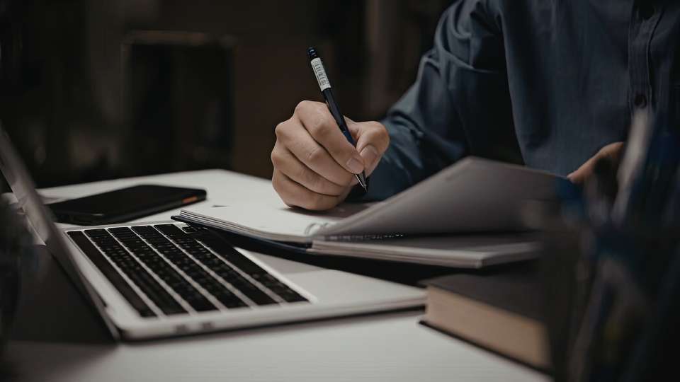 A close-up photograph of a person’s hands as they meticulously write in an open notebook with a blue pen. A laptop, a smartphone, and a thick book are also on the desk, lit by a soft, focused light that emphasizes the attention to detail. This image represents the careful, manual verification process required in the final stages of a project. It illustrates the text's advice that knowing exactly how to write co-author name in research paper is a critical, though often overlooked, step. By being precise with names and affiliations, researchers can avoid the accidental disrespect and resulting authorship disputes that arise from careless formatting errors.