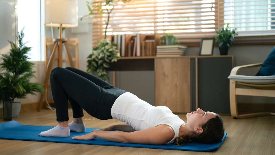 A woman performs a glute bridge on a blue mat, lifting her hips to activate her posterior chain. This exercise is an effective way to use home workout equipment for busy researchers to combat researcher back pain and improve posture.