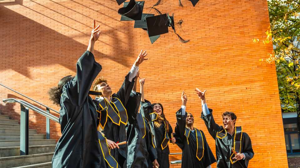 A group of graduates in black and gold robes toss their caps into the air in front of a brick building. This joyful moment represents the culmination of a long academic career, highlighting the importance of academic stamina and physical health in reaching such a milestone.