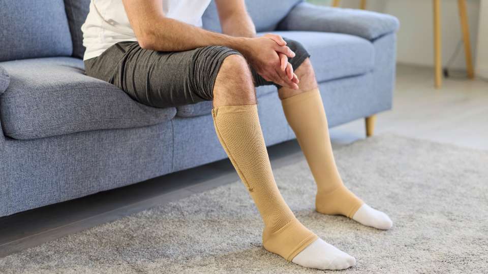 A person sits on a blue sofa wearing tan, zippered compression socks on their lower legs. This essential piece of travel workout gear helps researchers maintain circulation and energy levels during the demanding travel and networking hours of an academic career.