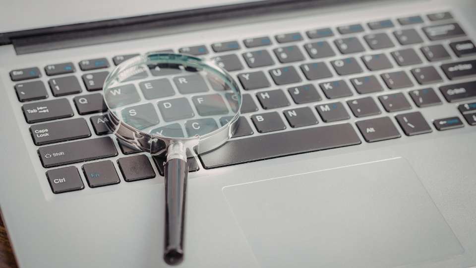 A magnifying glass lies on top of a silver laptop keyboard, symbolizing the intense scrutiny needed when evaluating international associates. This image illustrates the importance of background checks in an academic career, emphasizing the text's advice on how to accurately choose the right research partner or co-author to avoid unethical behavior and ensure a "drama-free" global collaboration.