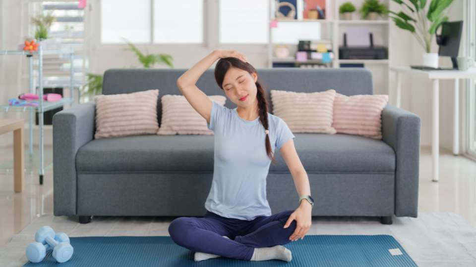 A woman sits cross-legged on a yoga mat in a living room, performing a gentle side neck stretch by tilting her head. This bedtime routine is a key recovery practice to alleviate tech neck symptoms and maintain the physical health required for high-level student performance.
