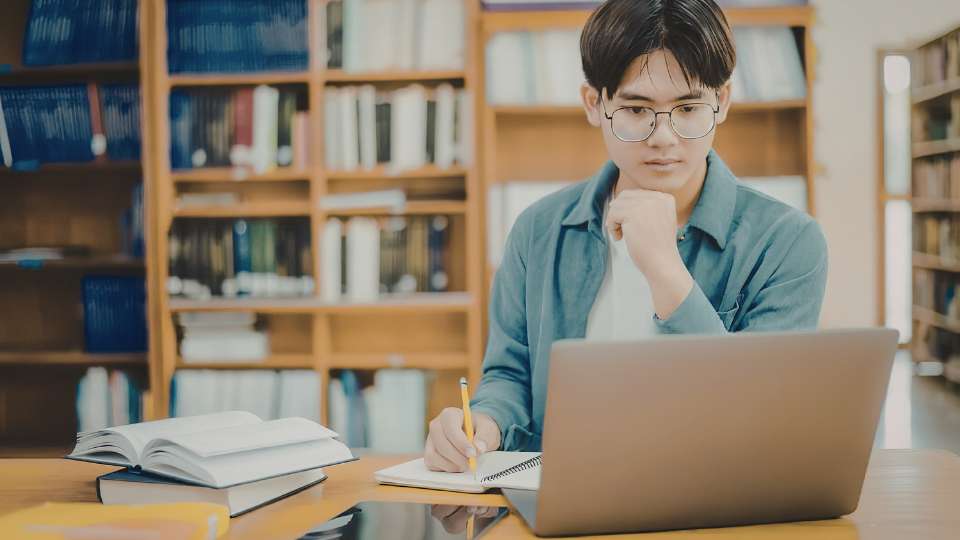 A young man with glasses and a denim shirt sits in a library, looking thoughtfully at his laptop screen with his hand on his chin. He is surrounded by bookshelves and has an open textbook and a notebook on the desk in front of him. This image represents the individual hard work and deep focus required in academic writing. It illustrates the text's warning about the "Phantom Contributor Problem," where a person who has not performed this level of labor may attempt to claim credit. By establishing clear criteria for what constitutes a primary writer, teams can avoid the frustration and authorship disputes that arise from casual reviewers demanding unearned status.