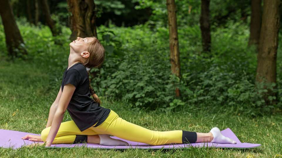 A girl performs a pigeon pose stretch on a yoga mat in a lush park setting. This movement represents the physical release necessary to combat researcher back pain and highlights the benefits of fitness in academic life for maintaining physical comfort.