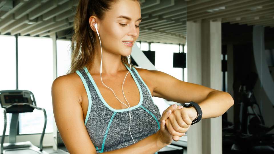 A woman in a gym checks her fitness tracker, symbolizing the use of the Pomodoro study technique to schedule short exercise breaks. These "movement snacks" are essential for building academic stamina and staying active while writing a thesis or manuscript.