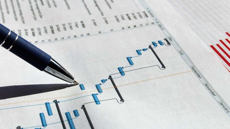 A close-up shot of a pen pointing to a detailed Gantt chart printed on paper, showing a series of interconnected project milestones across several months. This visual represents the "rock-solid project timeline" required to maintain accountability and strategic fund release throughout an academic career.