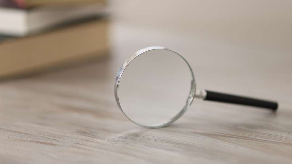 A magnifying glass lies on a wooden surface in front of a blurred stack of books. This image symbolizes the scrutiny required to detect predatory journals. It emphasizes the text's warning: researchers must use careful investigation to dodge scams that could permanently damage their academic career.