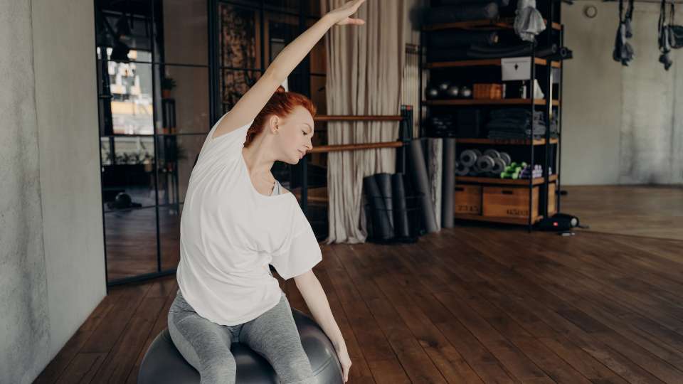 A woman with red hair performs side stretches while sitting on a stability ball in a studio. This image demonstrates how researchers can use active seating to strengthen their core and prevent the physical strain associated with tech neck symptoms during their academic career.