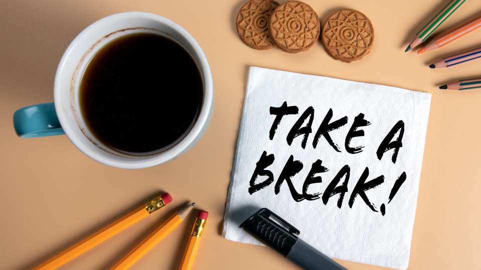 A top-down view of a coffee mug, cookies, pencils, and a napkin with "TAKE A BREAK!" written on it. This image emphasizes that rest is essential for maintaining academic stamina and ensuring successful long-term project management.