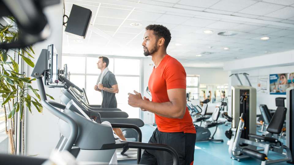 A man in an orange shirt runs on a treadmill in a bright, modern gym. This image illustrates how achieving small fitness targets builds the academic stamina and resilience needed to stay productive during a long research project.