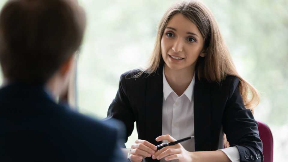 An over-the-shoulder view of a young woman with long brown hair wearing a dark professional blazer and a white collared shirt, listening and speaking during a serious ethical data collection interviews.