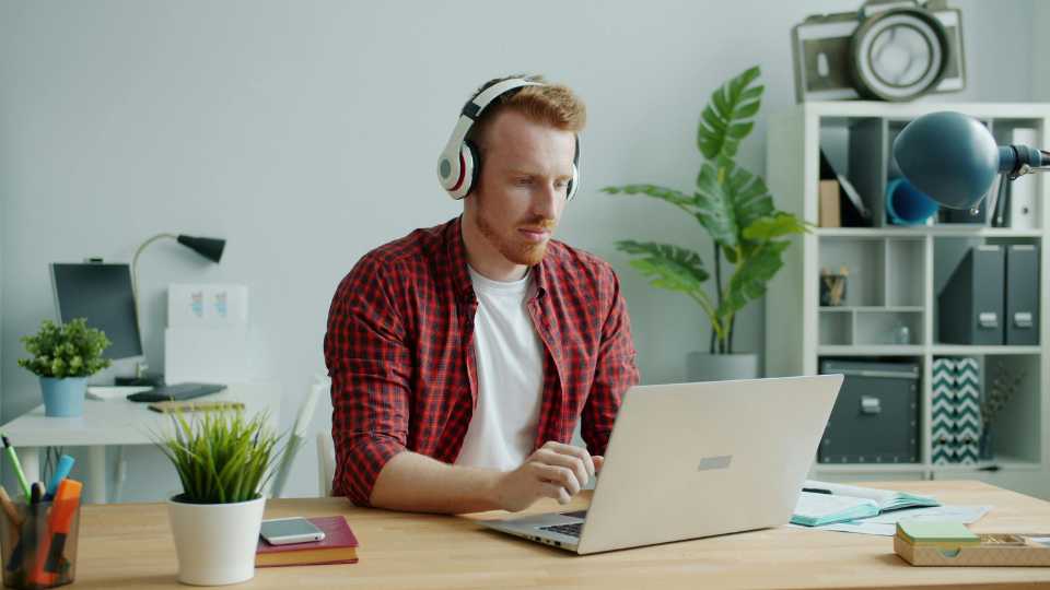 A red-haired man wearing a red plaid shirt and white headphones sits at a wooden desk, working intently on a silver laptop while transcribing or analyzing data in an office environment.