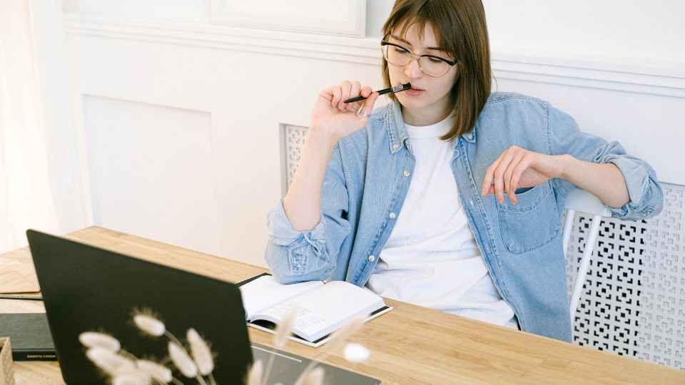 A thoughtful young woman with glasses and a denim shirt sits at a desk with a notebook and laptop, under the text "Use Autoethnography to Turn the Lens on Yourself".