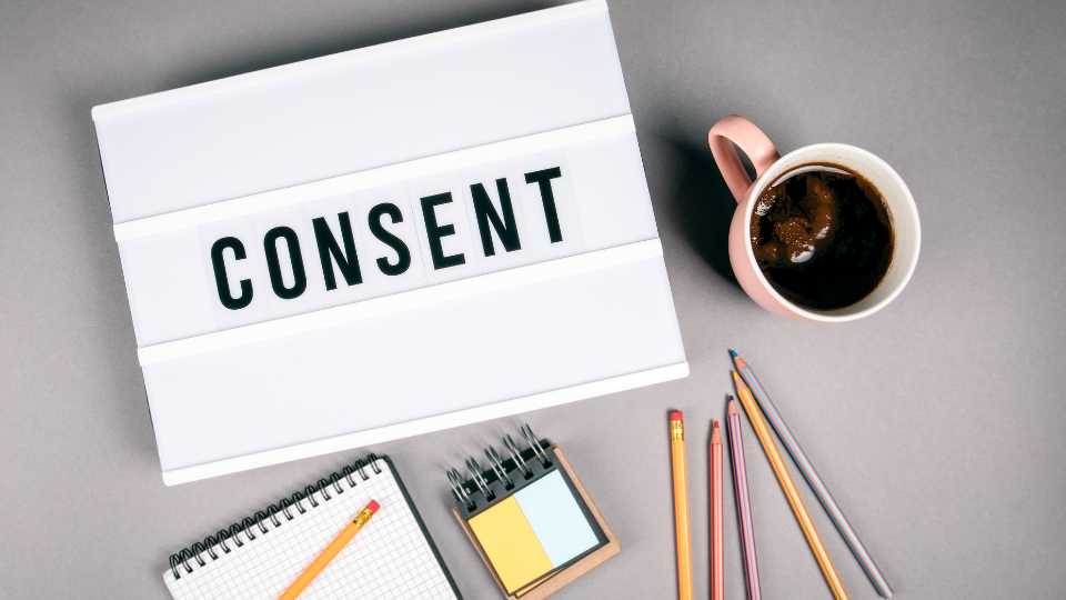 An overhead flat-lay shot on a grey background featuring a white light box displaying the word "CONSENT" in black capital letters, surrounded by a pink coffee mug, a spiral notebook, a small notepad, and several colored pencils.