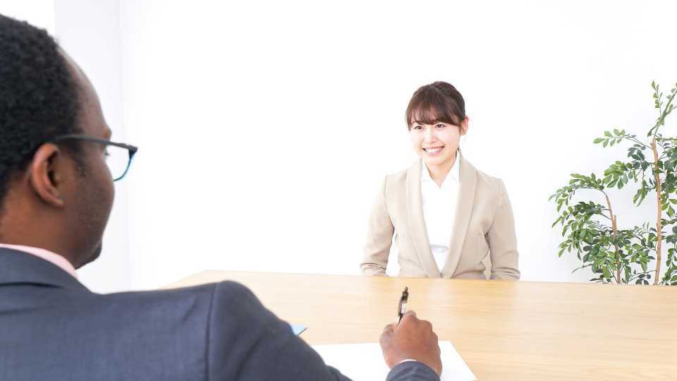 An over-the-shoulder view of a young woman with long brown hair wearing a dark professional blazer and a white collared shirt, listening and speaking during a serious ethical data collection interviews.