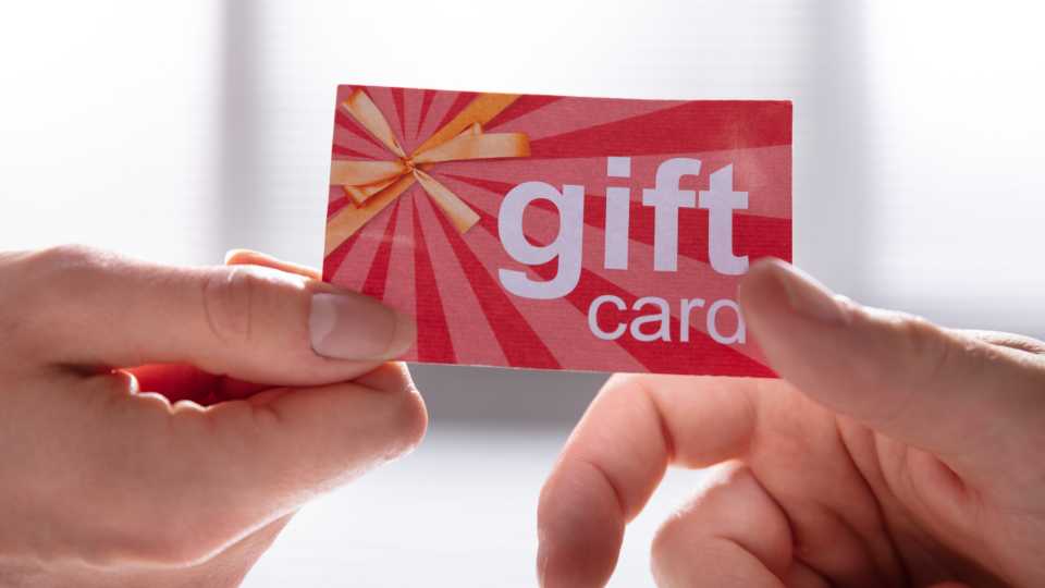 A close-up view of one person's hands handing a red, sunburst-patterned "gift card" with a gold ribbon graphic to another person's hands, representing the presentation of a participation incentive.