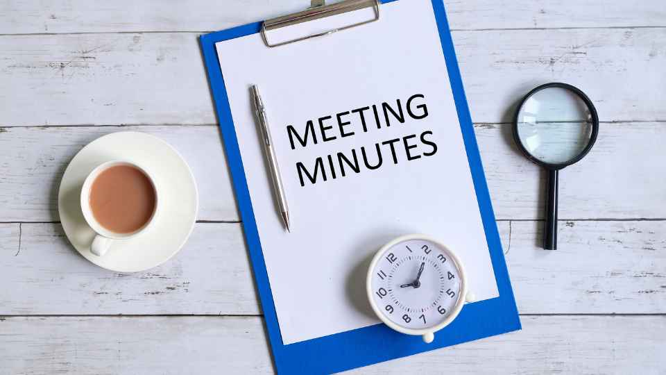 An overhead view of a blue clipboard holding a document titled "MEETING MINUTES" with a pen, positioned on a white wooden surface next to a cup of tea, a small white clock, and a magnifying glass.