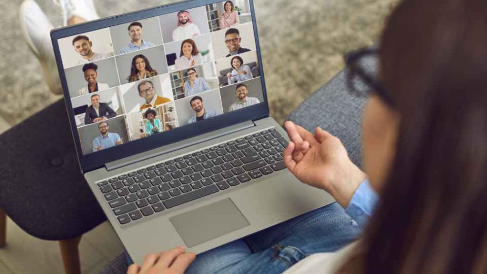 An over-the-shoulder view of a woman wearing glasses gesturing with her hand while hosting a large group video call on her laptop, showcasing multiple participant tiles on the screen.