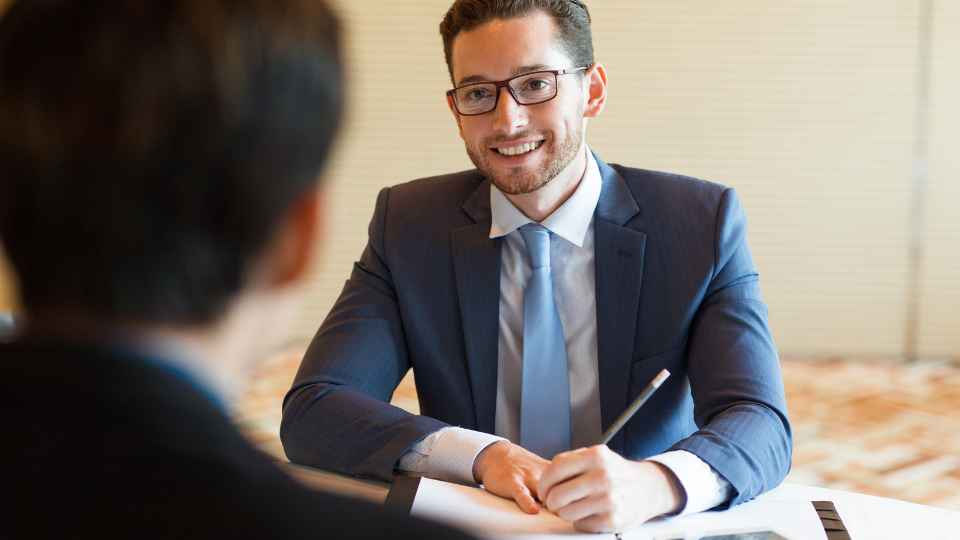 An over-the-shoulder view of a smiling young man with glasses and a beard, wearing a blue suit, light blue tie, and white shirt, engaged in a face-to-face interview or conversation.