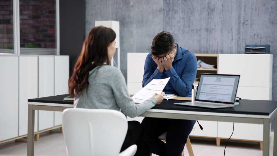 A man in a blue shirt sits at a desk across from a female interviewer, holding his face in his hands in a gesture of extreme stress or exhaustion while she reviews a document, illustrating a clear non-verbal cue that the session may be overwhelming.