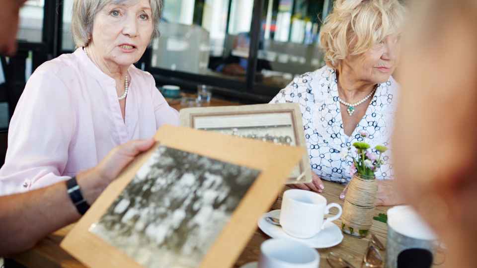 An over-the-shoulder view of an individual holding an old black-and-white photograph, sharing it with two older women sitting at a table with coffee cups, positioned under the header text "11. Use Photo-Elicitation to Spark Deeper Memories".