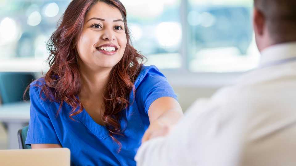 A close-up shot of a smiling Hispanic woman with reddish-brown hair wearing blue medical scrubs shaking hands with a person in a white shirt, illustrating the process of establishing trust and rapport with a participant.