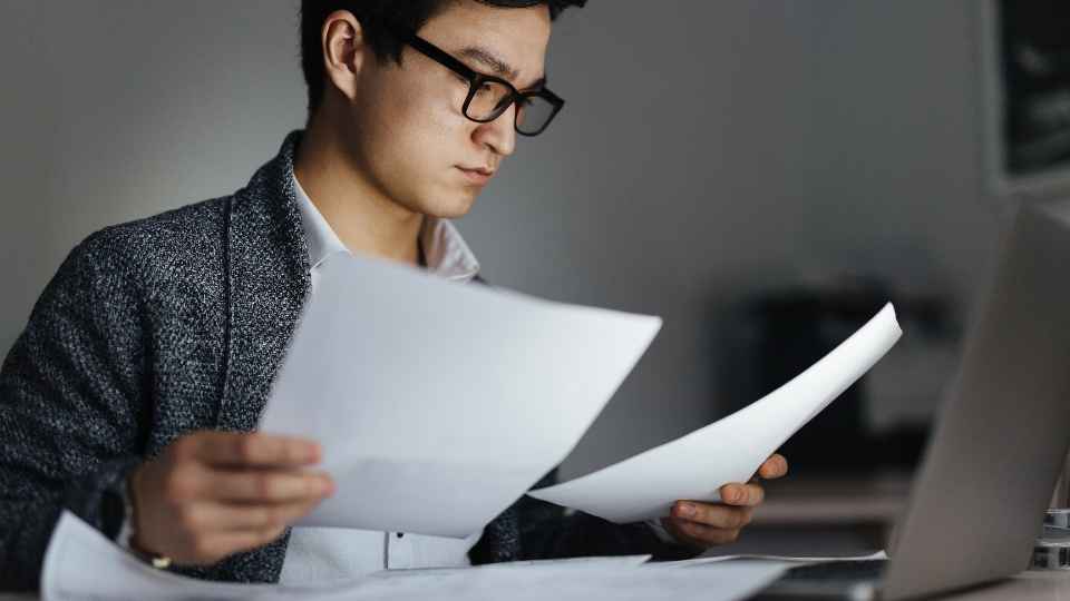 A young man wearing glasses and a grey tweed jacket sits at his desk, focused intently on reviewing printed documents held in his hands next to a laptop, positioned below the heading text "6. Read Between the Lines for Silent Gaps".