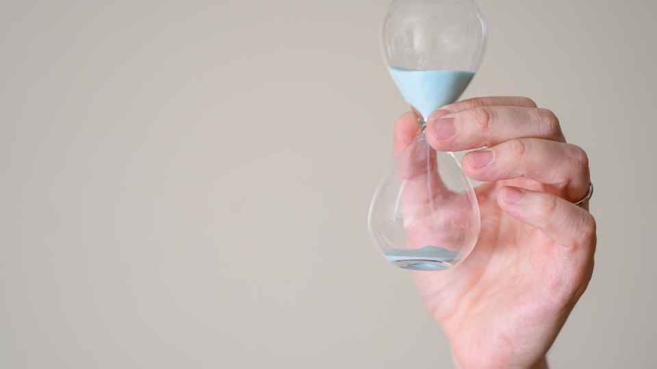 A close-up shot of a hand holding a clear glass hourglass with light blue sand running through it against a plain off-white background, representing the implementation of time limits in a focus group discussions protocol.