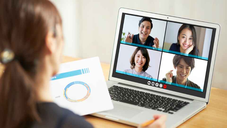 An over-the-shoulder view of a woman holding a document with a circular chart while looking at a laptop screen that shows a four-way video conference call with smiling participants.